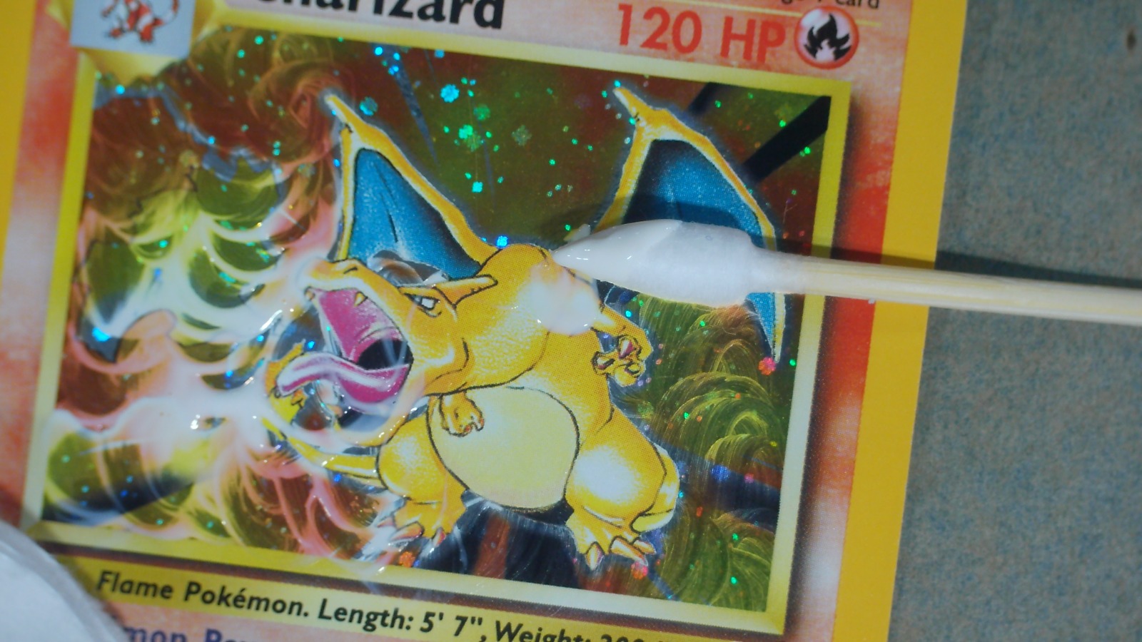 Q-tip cotton swab applying cleaning solution to a holographic Charizard Base Set 2 Pokemon card during Apex surface prep